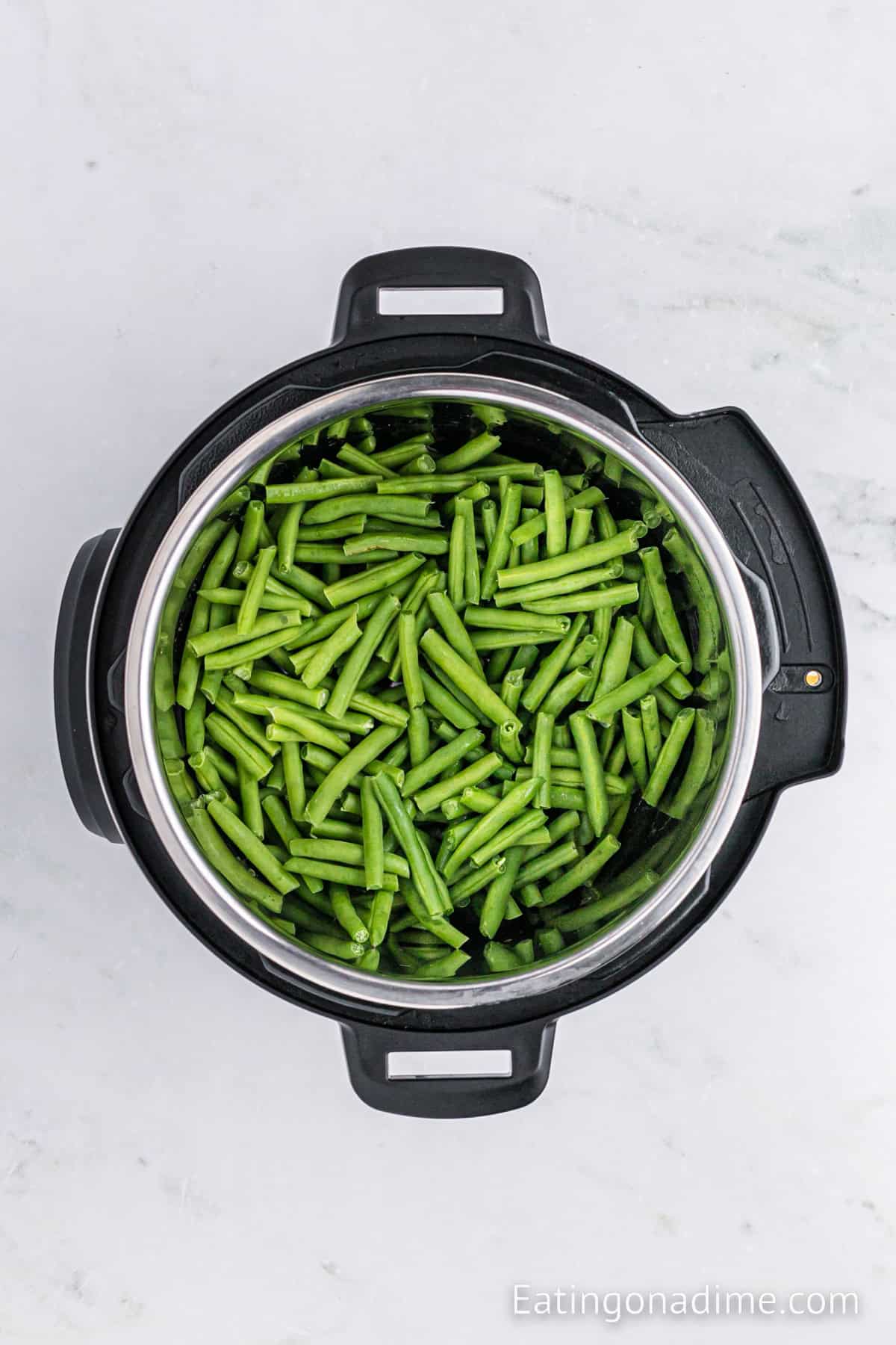 An overhead view of an Instant Pot Green Bean Casserole, featuring fresh, cut green beans. The Instant Pot is placed on a light-colored marble countertop. The website "Eatingonadime.com" is watermarked in the lower right corner of the image.