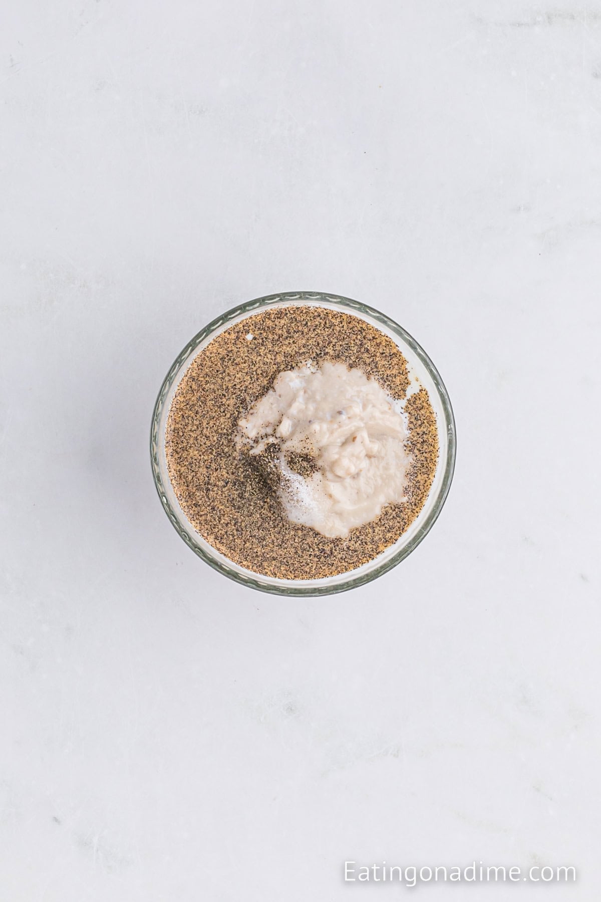 A glass container filled with a mixture of ground black pepper and a whitish, creamy substance, placed on a light gray, marble-textured surface. The website name "Eatingonadime.com" is visible in the bottom right corner, possibly showcasing ingredients for an Instant Pot Green Bean Casserole.