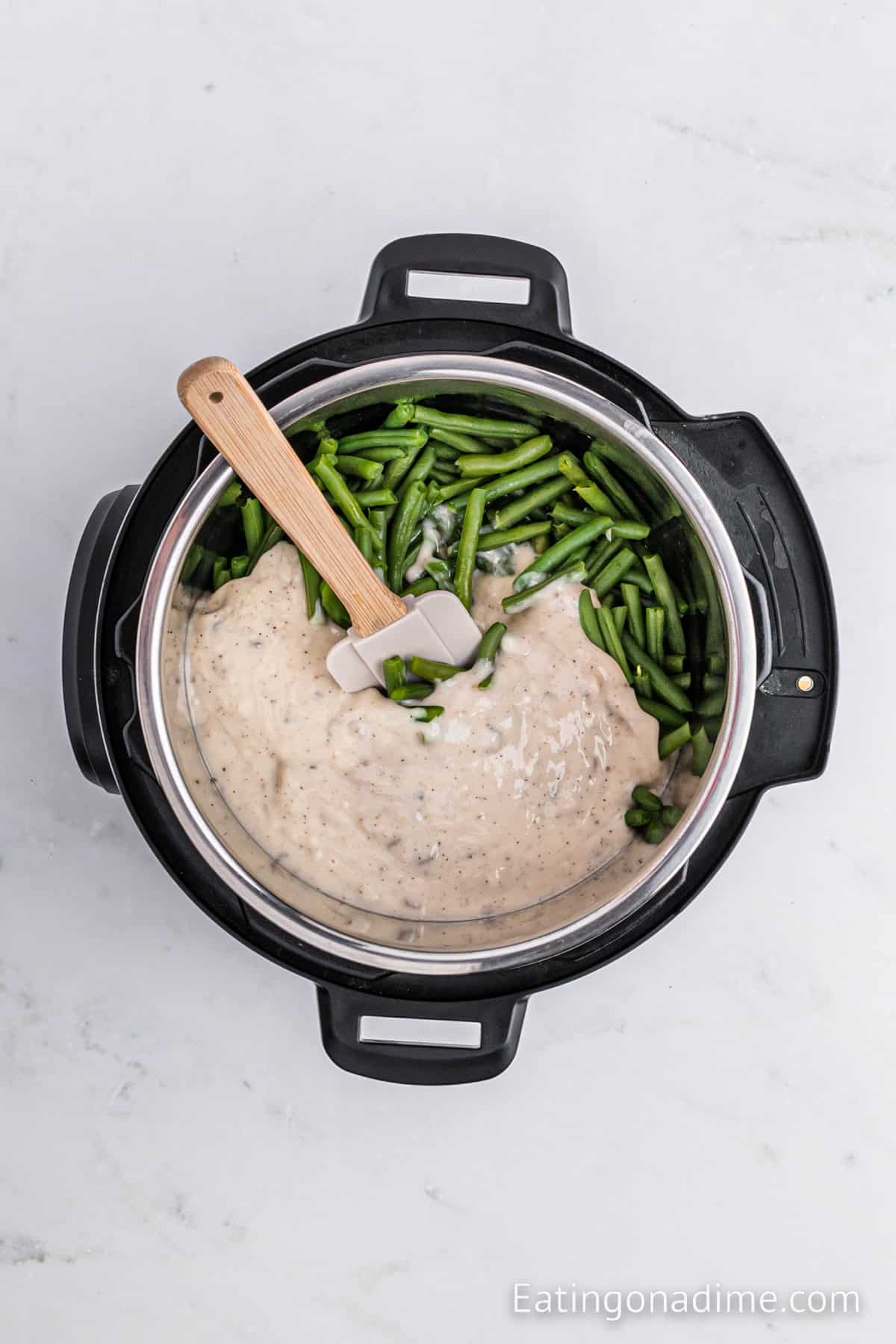 An open Instant Pot is shown from above on a white surface. Inside the pot, there are fresh green beans and a creamy, thick white sauce being mixed with a white spatula—perfect for an Instant Pot Green Bean Casserole. The website name, Eatingonadime.com, is visible in the bottom right corner of the image.