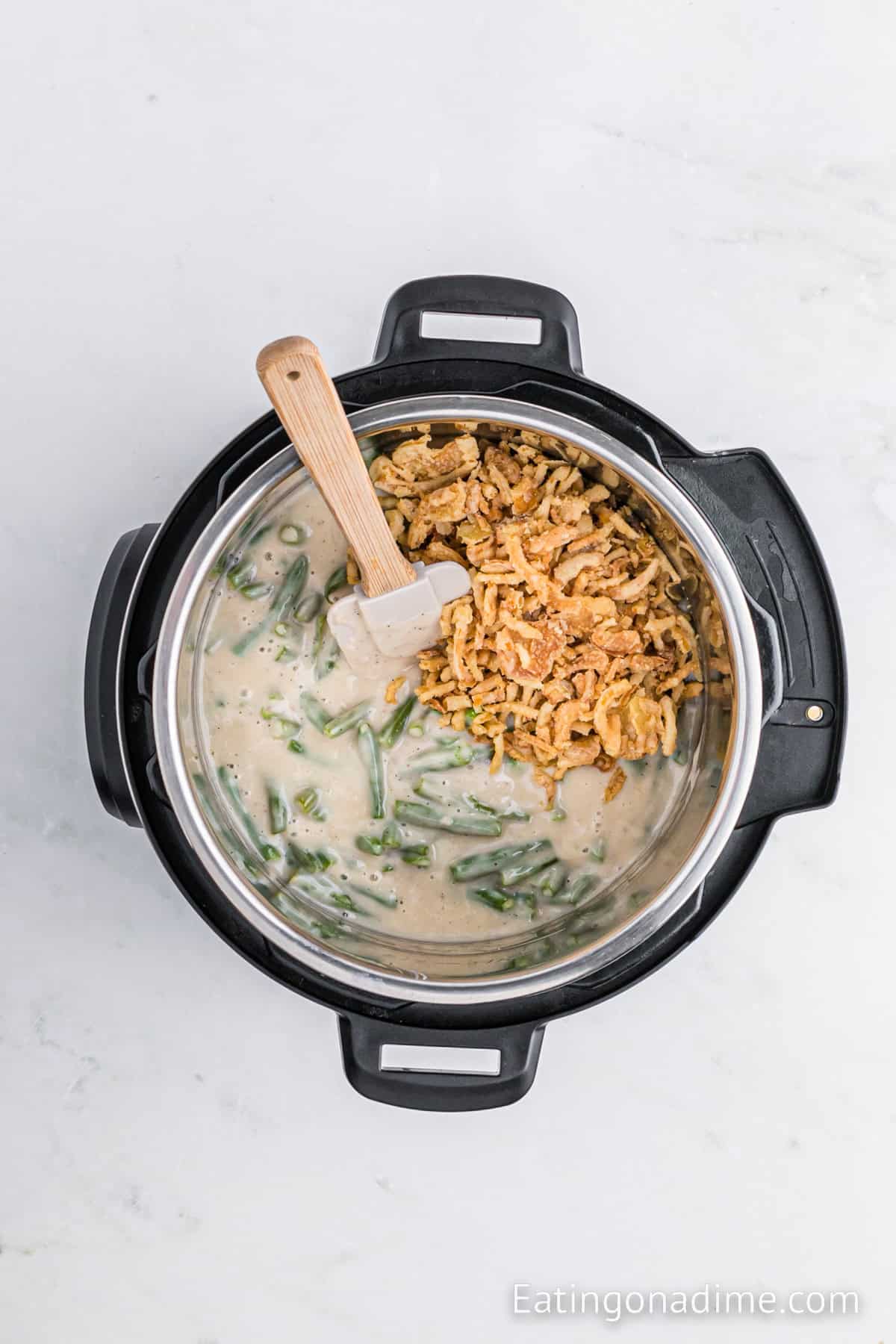 A top view of an Instant Pot Green Bean Casserole reveals a creamy green bean mixture, topped with crispy fried onions. A spatula rests on the edge of the cooker, and the background is a light marble surface.