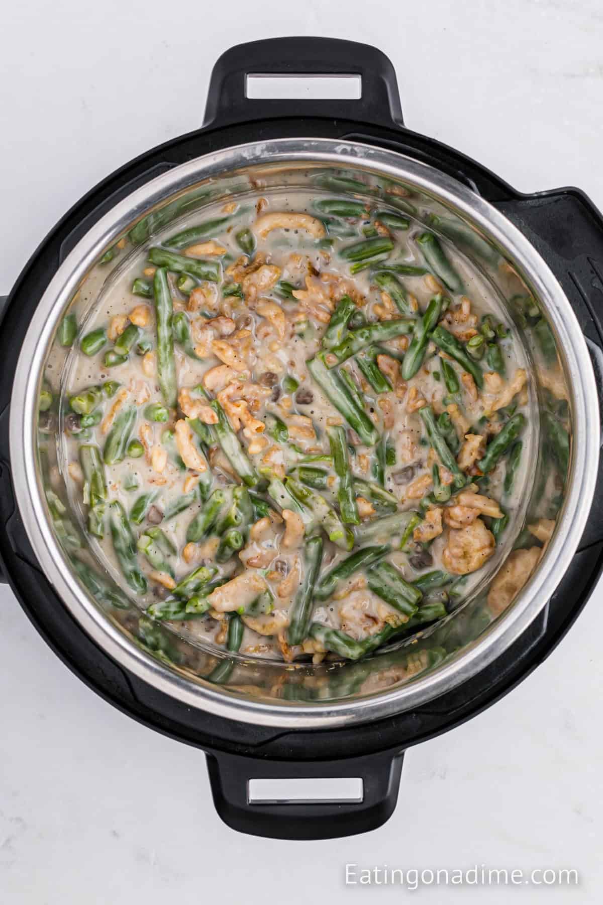 An instant pot filled with a creamy dish consisting of cooked ground meat and green beans, topped with a sauce. This Instant Pot Green Bean Casserole appears well-cooked and seasoned, and the appliance is situated on a white surface with text reading "Eatingonadime.com" in the corner.