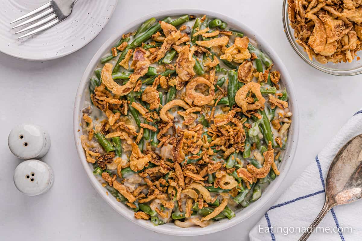 A bowl of Instant Pot Green Bean Casserole topped with crispy fried onions, placed next to a dish with additional fried onions, a salt and pepper shaker, a fork on a partial plate, and a cloth napkin with a spoon on it. The dish looks fresh and delicious.