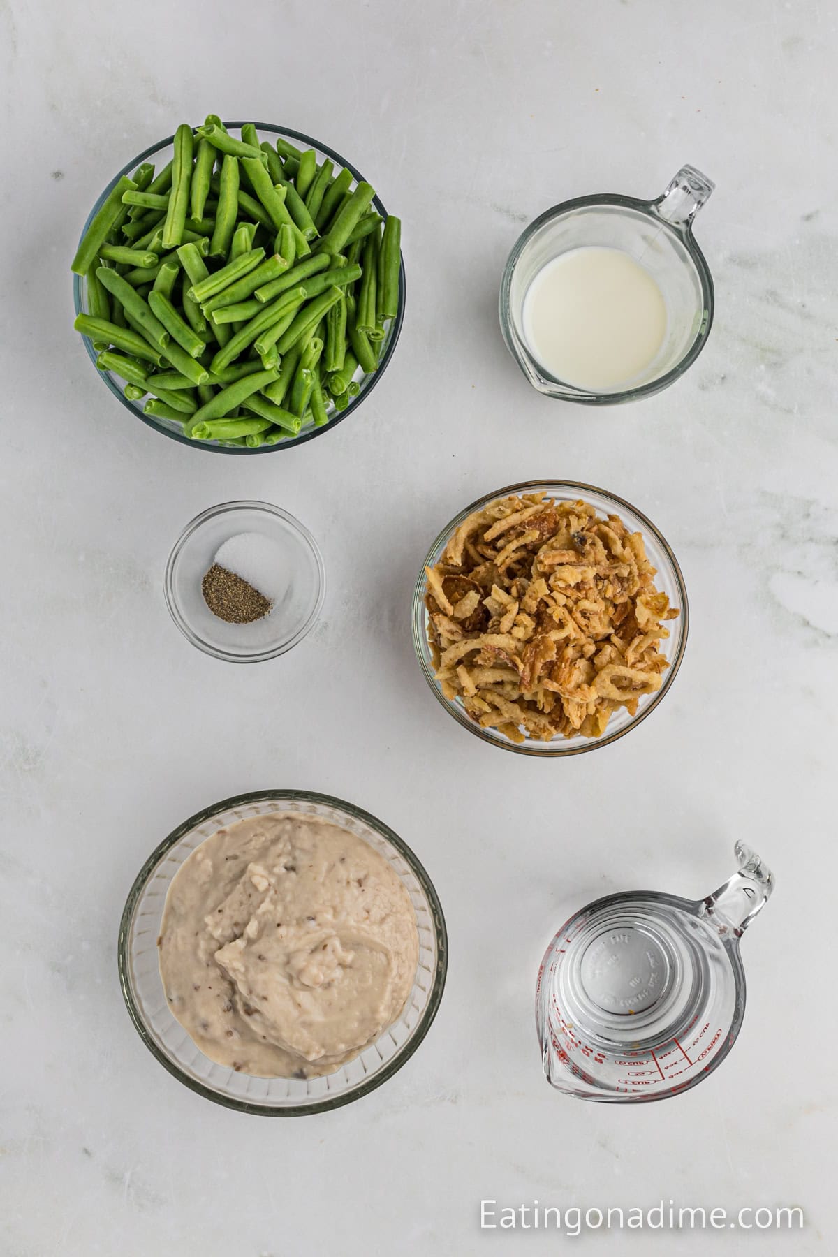 Top-down view of ingredients for an Instant Pot Green Bean Casserole, including a bowl of green beans, a bowl of crispy fried onions, a bowl of creamy mushroom soup, a measuring cup of milk, a small bowl of black pepper, and a measuring cup of water on a light surface.