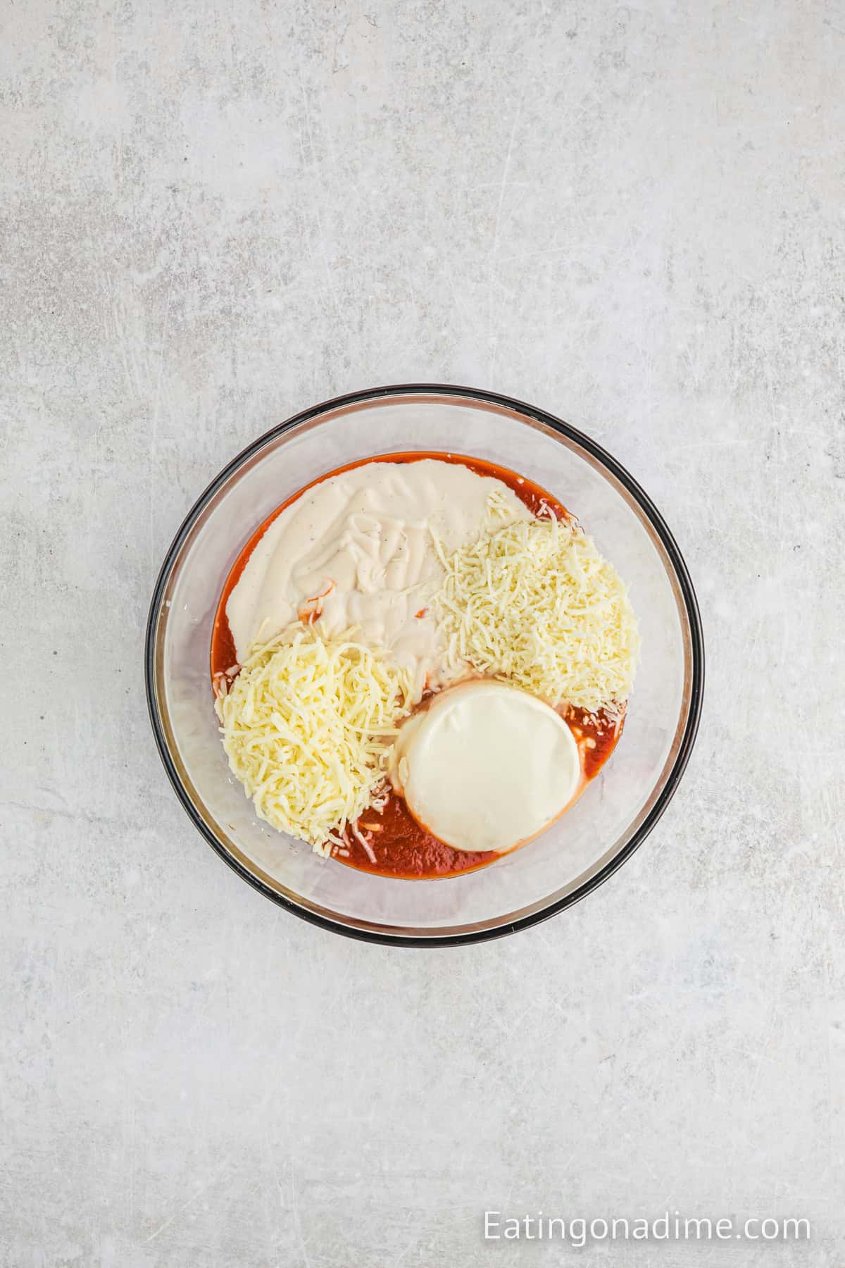 A glass bowl holds layers reminiscent of Olive Garden's 5 Cheese Ziti, with grated white cheese, red sauce, a round block of cream cheese, and a creamy mixture. It's all set against a light textured background.