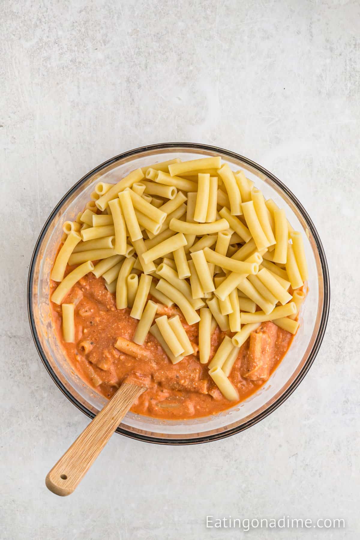 A glass bowl filled with cooked pasta sits atop a luscious, creamy red sauce reminiscent of Olive Garden's 5 Cheese Ziti. A wooden spoon rests in the bowl, partially immersed in the savory delight. The scene unfolds on a white, textured surface.