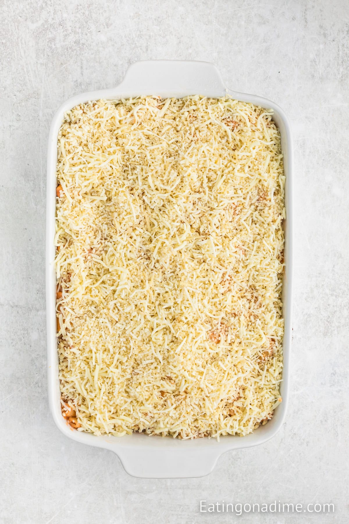 A white casserole dish brimming with an unbaked creamy chicken Olive Garden 5 Cheese ziti, topped with shredded cheese and breadcrumbs, rests on a light gray surface.