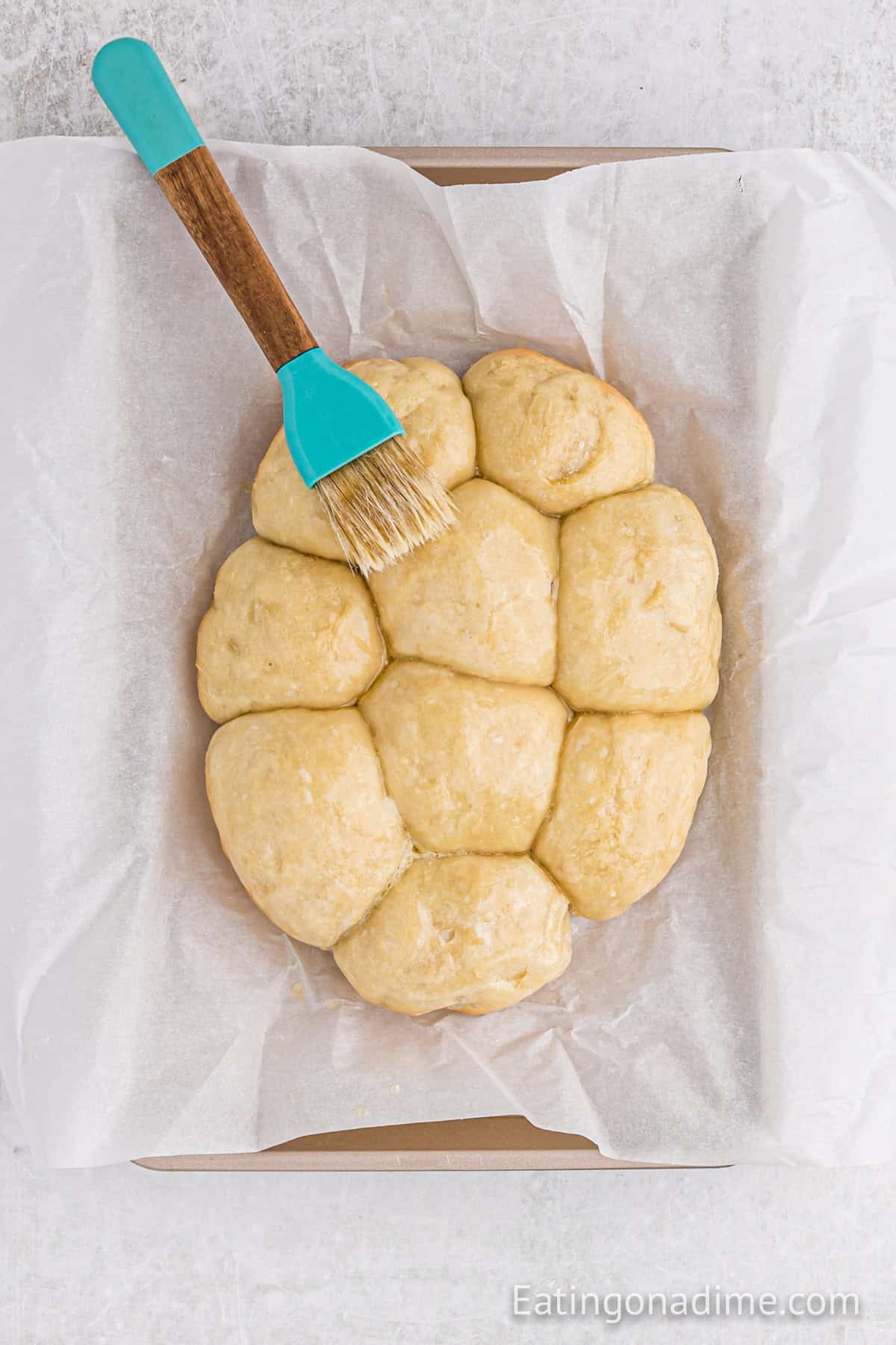 Brushing melted butter with dinner rolls on a baking sheet lined with parchment paper