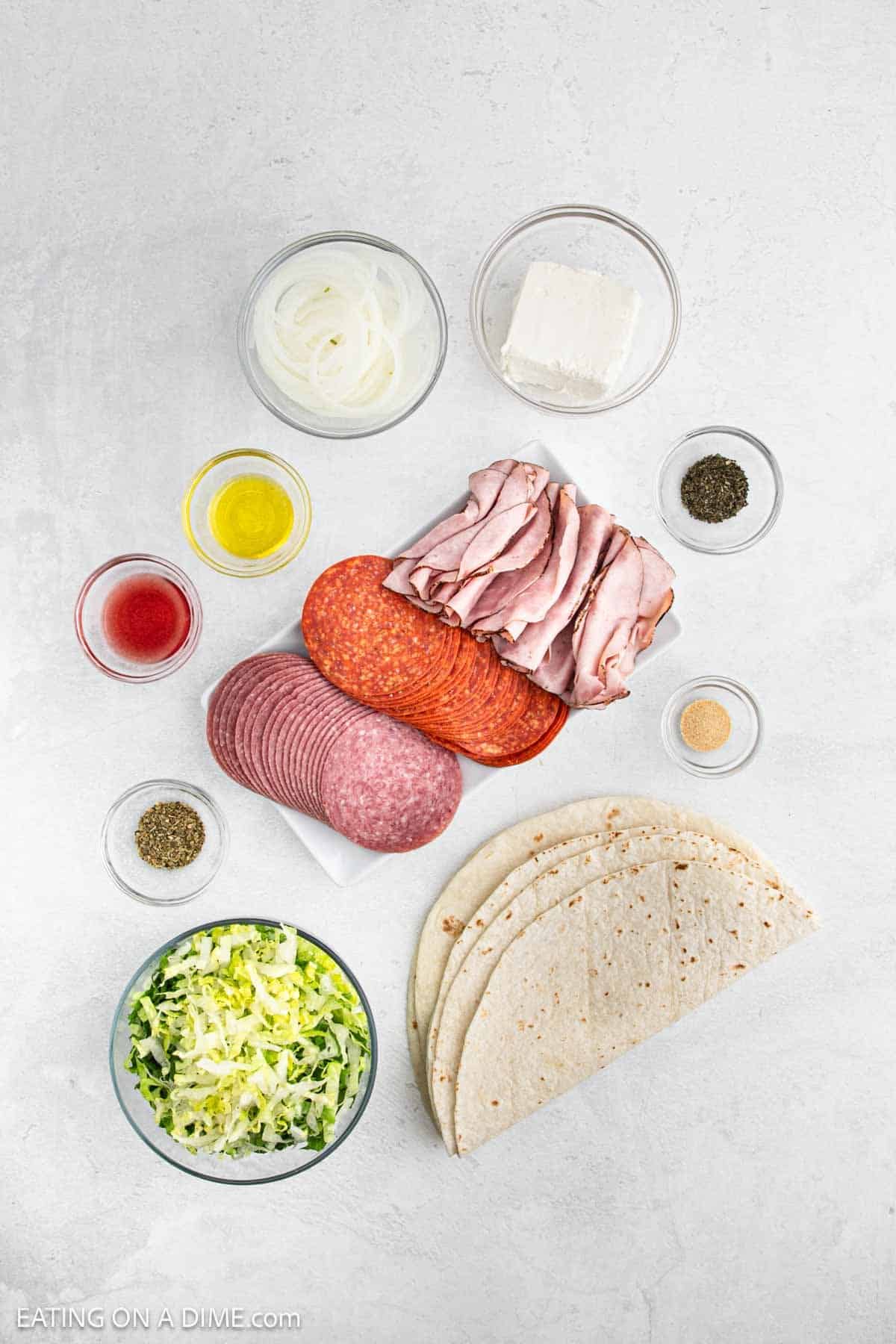 Flat lay of wrap ingredients for pinwheel sandwiches on a white surface: tortillas, shredded lettuce, sliced onions, cream cheese, pepperoni, roast beef, salami, and small bowls of oil, vinegar, black pepper, garlic powder, and seasoning.