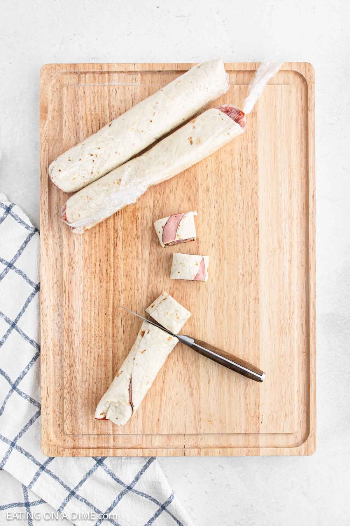 A wooden cutting board displays two whole rolled tortillas, one partially sliced into pinwheel sandwiches. A knife rests beside the neatly sliced pieces, while a checkered cloth peeks in from the left.