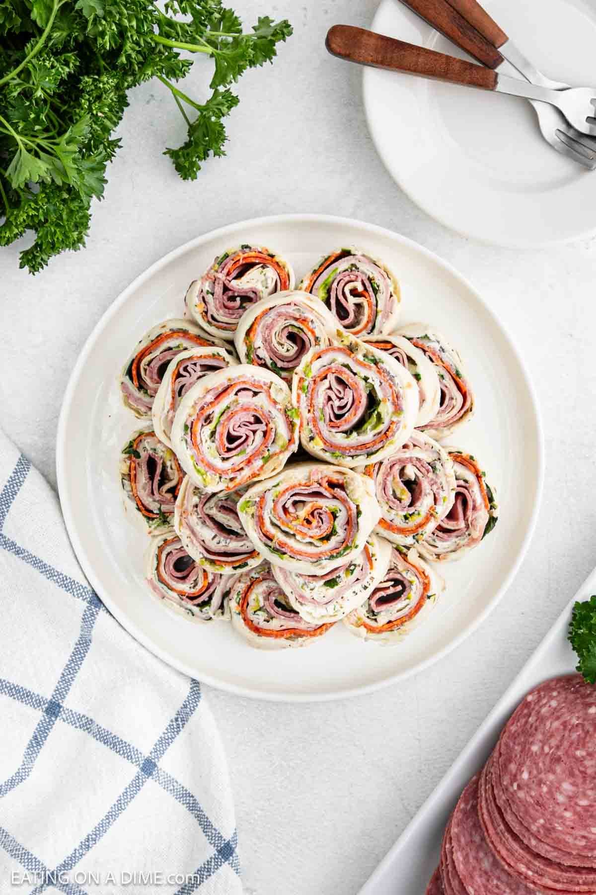 A plate of pinwheel sandwiches filled with sliced meat, cheese, and spinach, arranged in a spiral pattern. Parsley garnish is nearby, along with a stack of white plates and forks. A checkered cloth partially visible on the left completes the inviting setup.