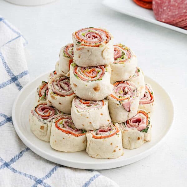 A white plate stacked with delicious pinwheel sandwiches made from rolled flatbreads filled with slices of deli meat, visible seasoning, and fresh greens. The plate is set on a white surface beside a blue and white checkered cloth.