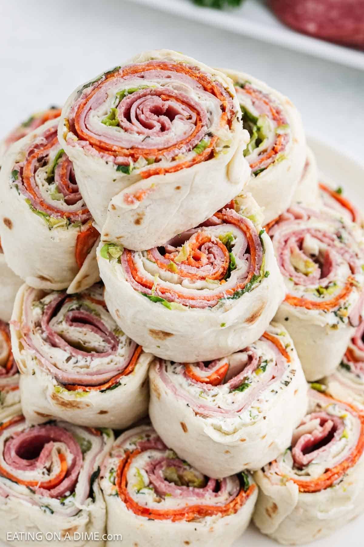 A stack of pinwheel sandwiches made with tortilla wraps, layered with slices of meat, lettuce, cheese, and vegetables. These pinwheel sandwiches are neatly arranged on a plate, showcasing colorful spiral patterns.