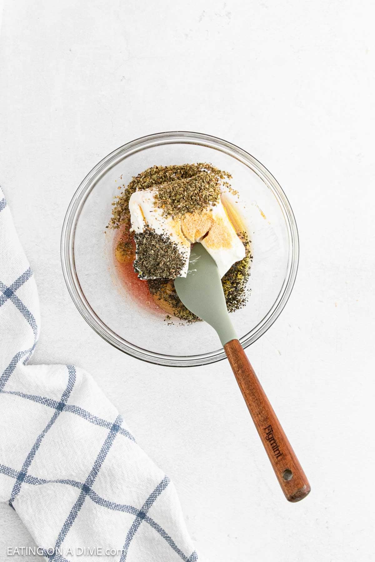 A glass bowl contains cream cheese, herbs, and spices, ready to be mixed for delightful pinwheel sandwiches. The bowl is placed on a white surface next to a blue and white checkered cloth.