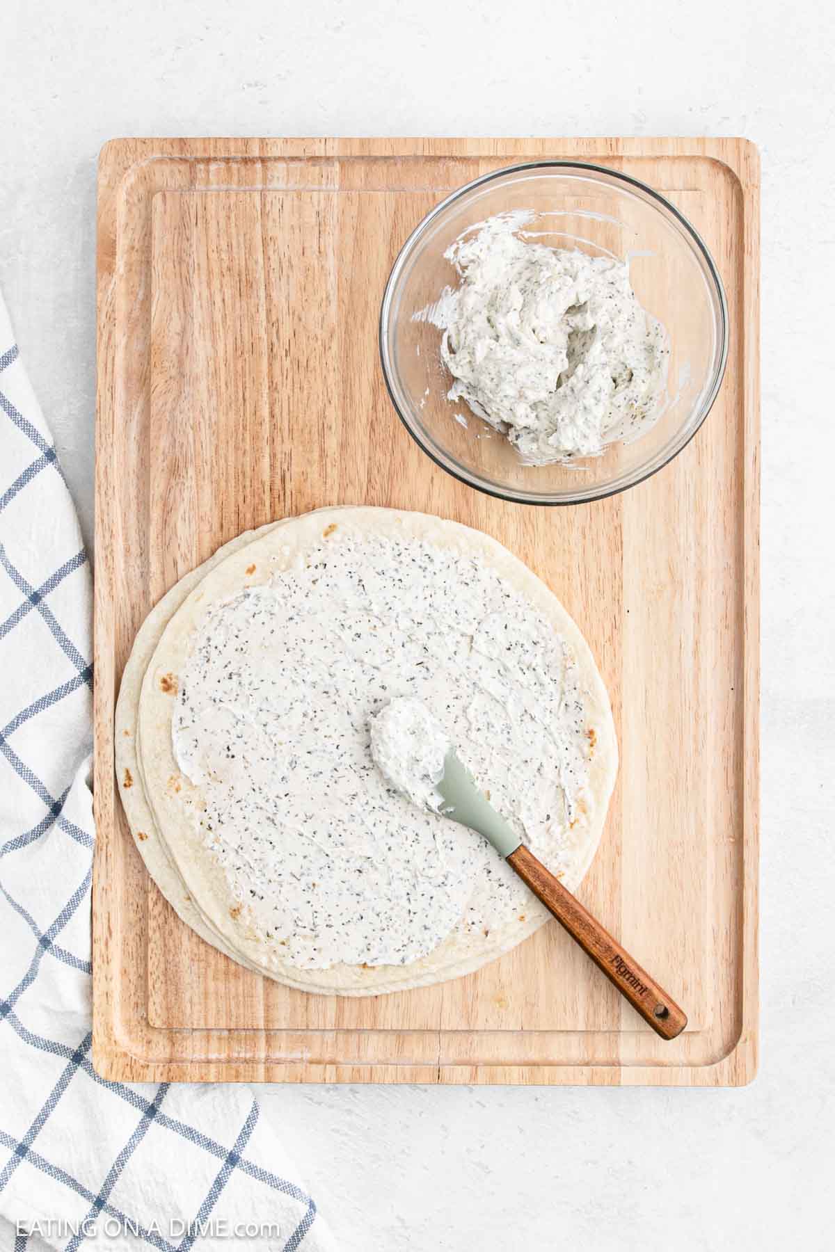 A wooden cutting board showcases pinwheel sandwiches made from tortillas spread with herb cream cheese. A spatula rests on one tortilla, ready to roll. Nearby, a glass bowl of cream cheese sits invitingly, while a blue and white checkered cloth peeks in from the left.