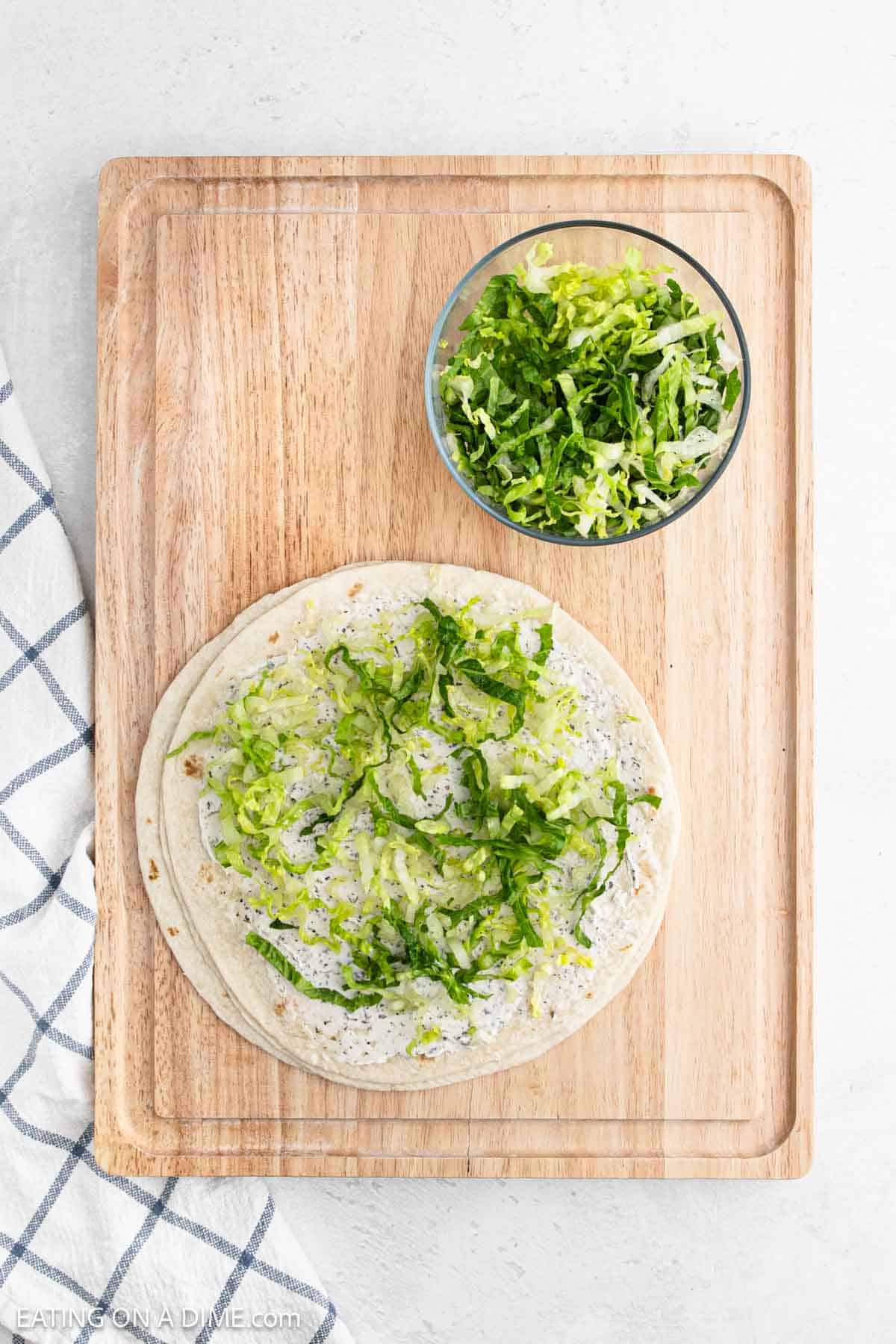 A flatbread topped with a creamy spread and shredded lettuce rests on a wooden cutting board, resembling delectable pinwheel sandwiches. A bowl of extra shredded lettuce sits beside it, while a white and blue checkered cloth peeks out from the side.