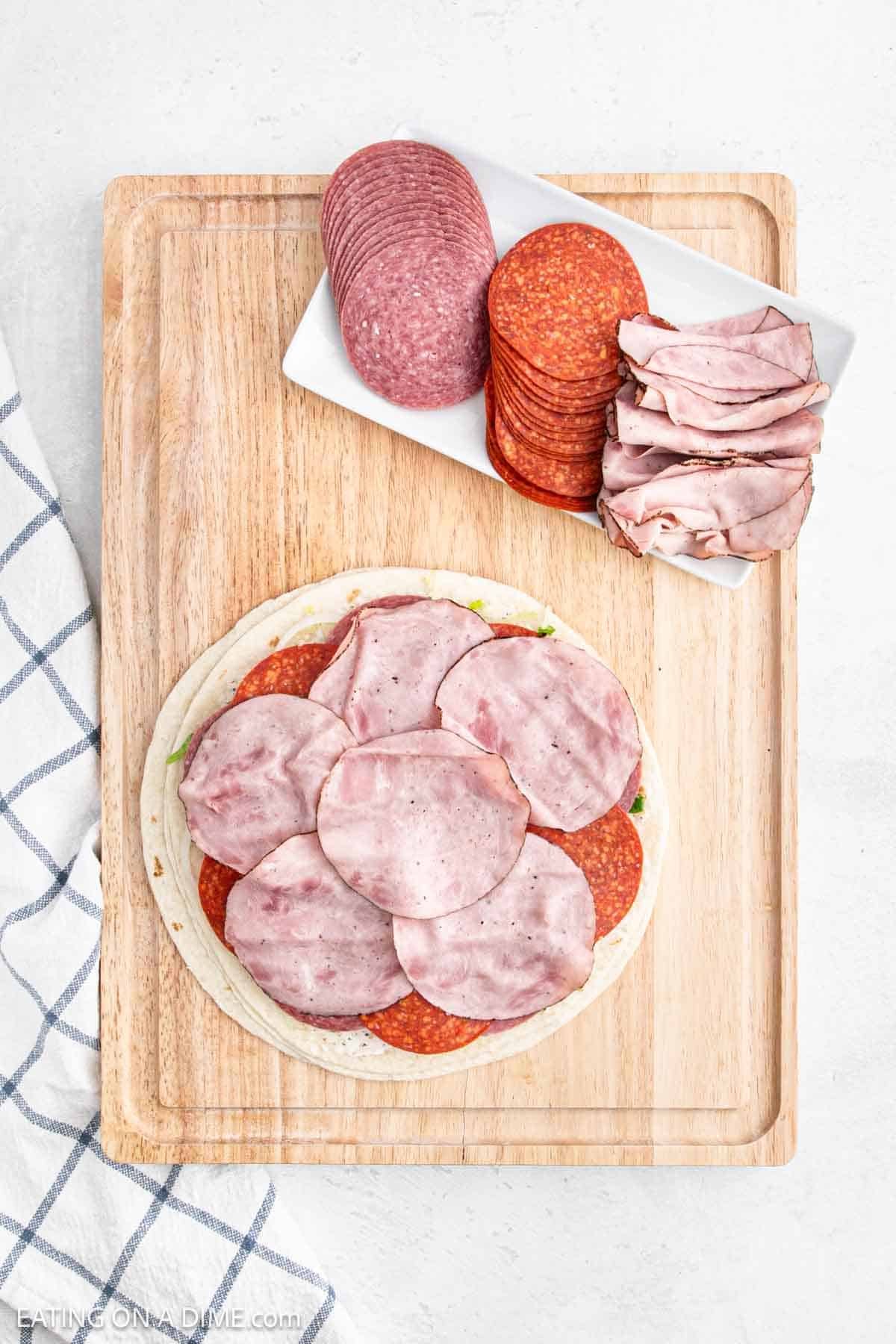 A wooden board displays an open-faced sandwich topped with slices of ham and pepperoni, reminiscent of pinwheel sandwiches. Nearby, a white dish holds additional slices of salami, pepperoni, and ham. A checkered cloth is partially visible on the side.