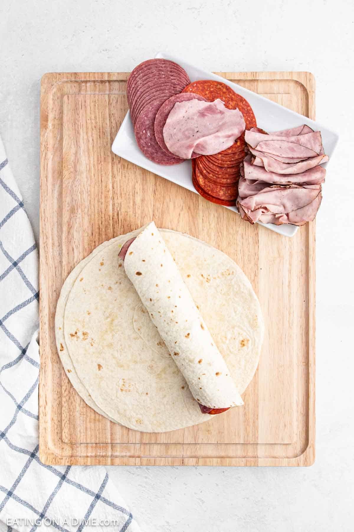 A partially rolled tortilla, reminiscent of pinwheel sandwiches, with deli meats inside is placed on a wooden cutting board. Beside it, a small rectangular white plate holds stacked slices of salami, pepperoni, and roast beef. A striped cloth napkin is visible to the side.