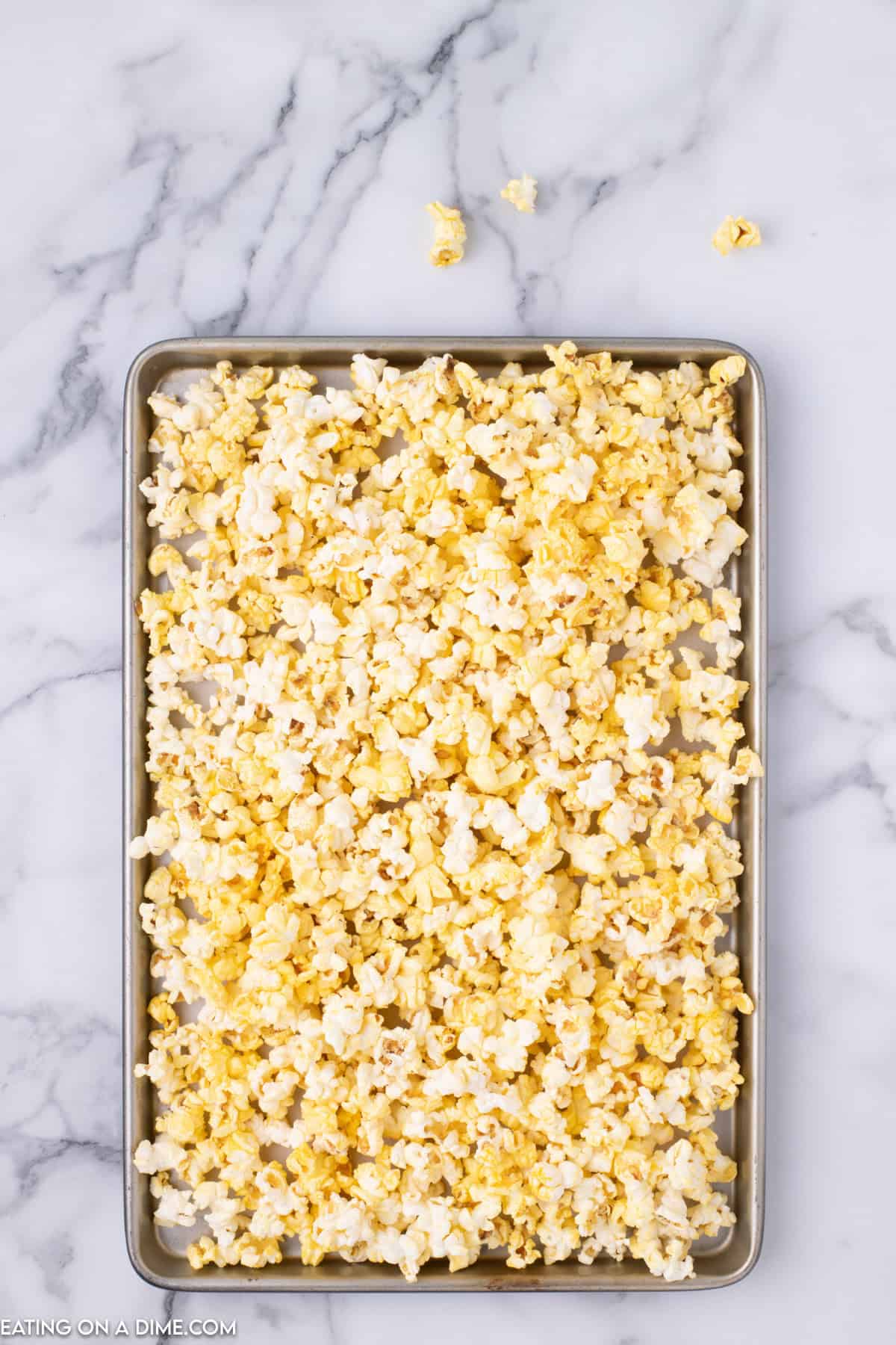 A baking sheet filled with a large amount of freshly popped Christmas Crunch popcorn sits on a marble countertop. A few scattered pieces of popcorn lie around the edges of the tray. The festive mix appears evenly buttered and has a light golden color.