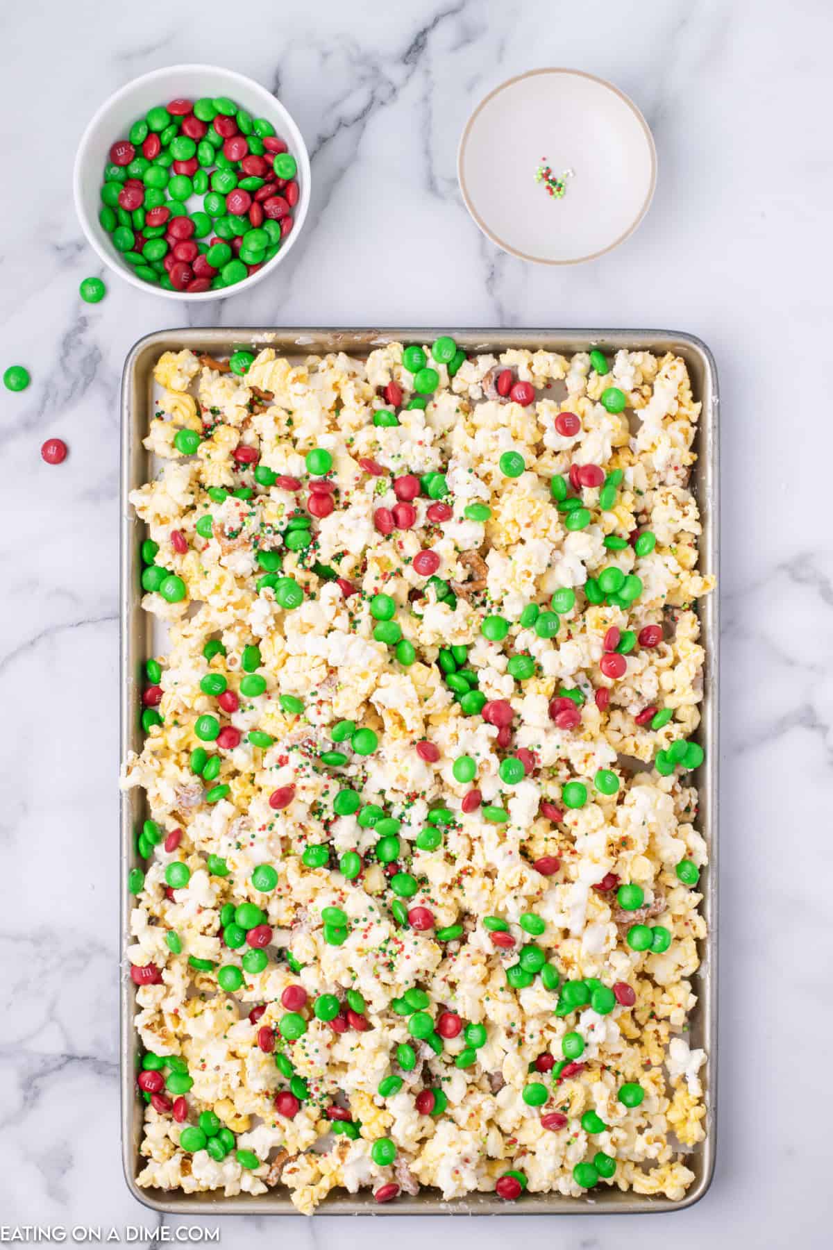 A tray of Christmas Crunch, popcorn mixed with red and green M&M candies, sits on a marble countertop. Nearby, a small bowl contains extra M&Ms, while another bowl has a few scattered sprinkles. The tray is filled to the rim with the festive popcorn mixture.