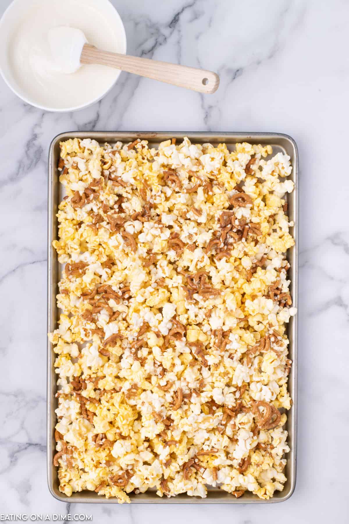 A baking sheet filled with freshly made caramel corn sits on a marble countertop, looking perfect for a batch of Christmas Crunch. A small bowl with a white spatula, filled with melted white chocolate, is placed in the top left corner of the image.