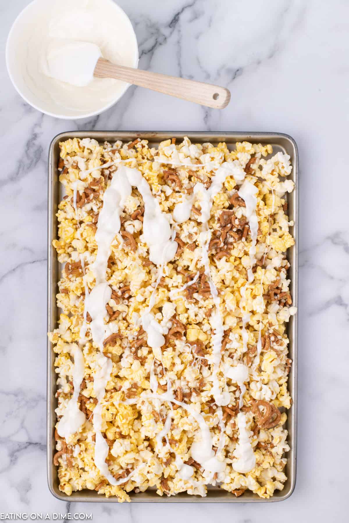 A tray of Christmas Crunch caramel popcorn drizzled with white chocolate, placed on a white marble surface. A small bowl of melted white chocolate with a wooden spatula is near the top-left corner.