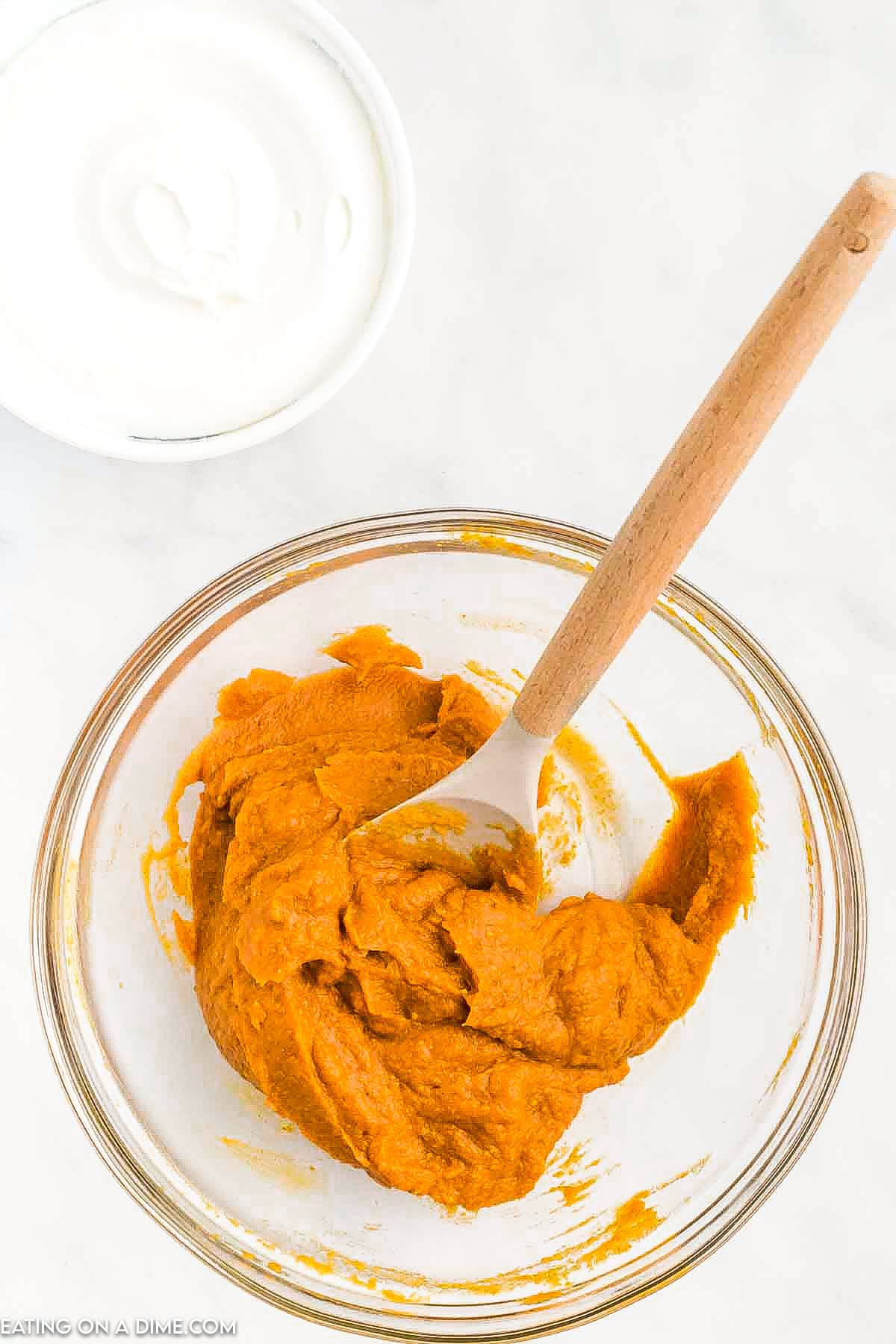 A bowl of pumpkin puree with a wooden spoon rests on a white surface, ready for your no bake pumpkin pie recipe. Nearby, there is a small bowl of creamy yogurt.