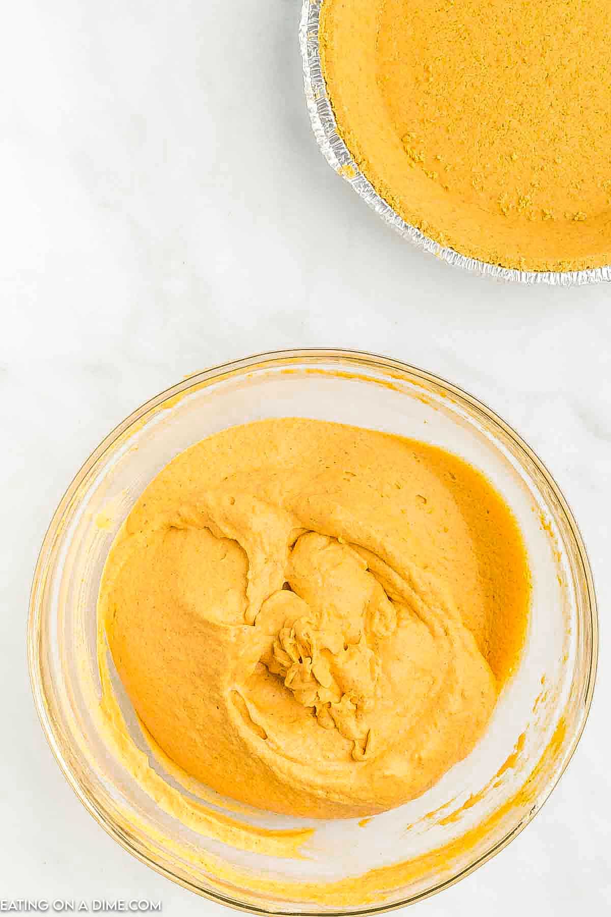 A glass bowl filled with creamy pumpkin pie filling sits on a marble surface, perfect for a quick no-bake pumpkin pie recipe. Nearby, there's a partially visible foil pie crust shell, ready to be filled with the luscious pumpkin mixture.