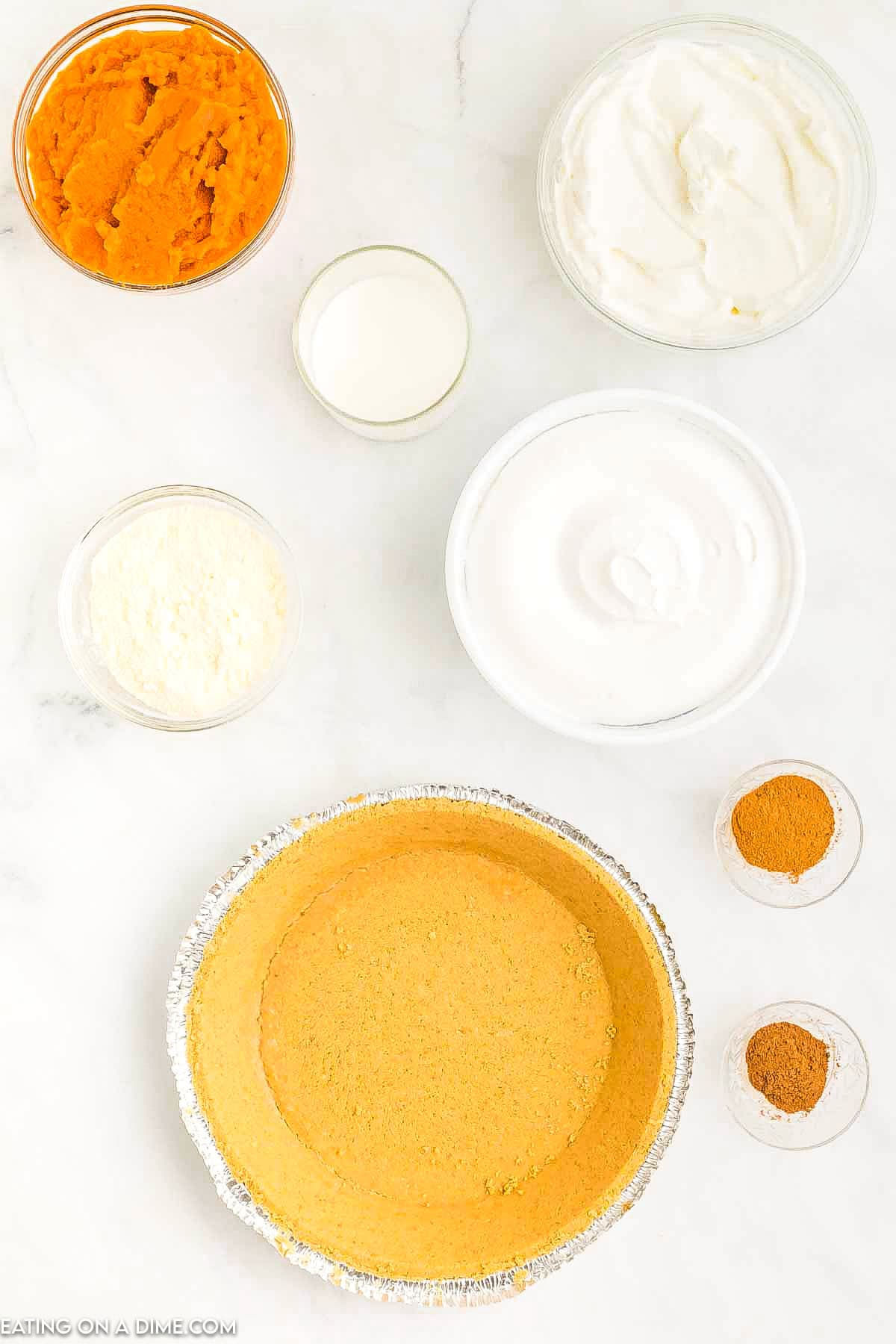 Top-down view of ingredients for a No Bake Pumpkin Pie Recipe: a graham cracker crust, bowls of pumpkin puree, whipped topping, cream cheese, condensed milk, and small bowls of cinnamon and nutmeg on a light surface.