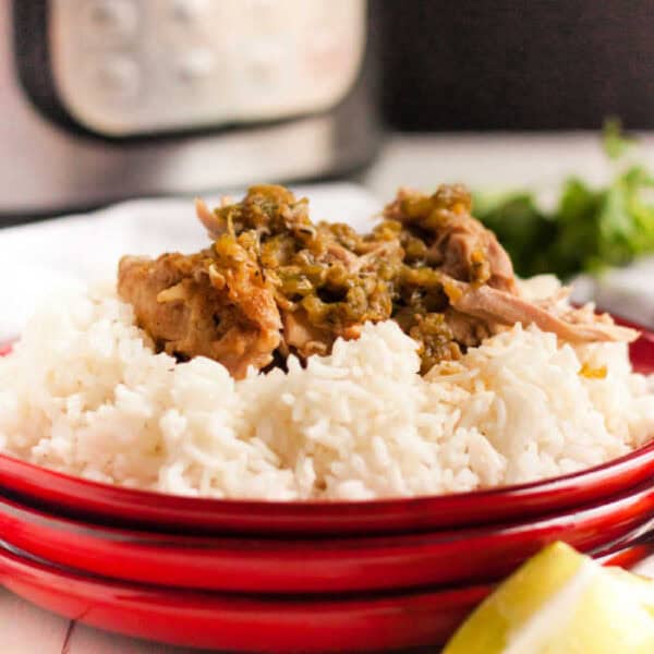 A red plate holds a serving of white rice topped with shredded pork and green salsa, reminiscent of a zesty Instant Pot Salsa Verde Chicken recipe. In the background, there's a blurred pressure cooker and some cilantro. A lime wedge is positioned in the corner of the image.