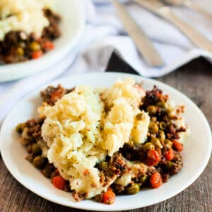 A plate of shepherd's pie, crafted from a delectable crock pot recipe, features a golden layer of mashed potatoes atop a savory blend of ground meat, peas, and carrots. Nestled on a wooden table, the dish is accompanied by napkin and cutlery in the background.