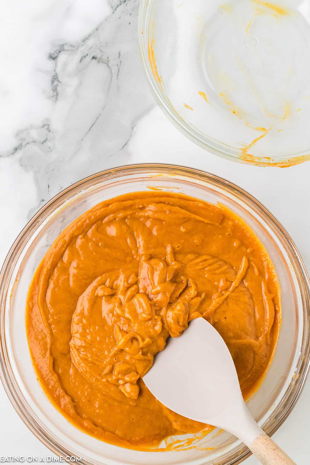 A glass bowl filled with creamy pumpkin batter, reminiscent of a Starbucks Pumpkin Bread recipe, sits on a marble surface. A white spatula rests inside the bowl, partially covered in batter. Another empty glass bowl is visible in the corner.