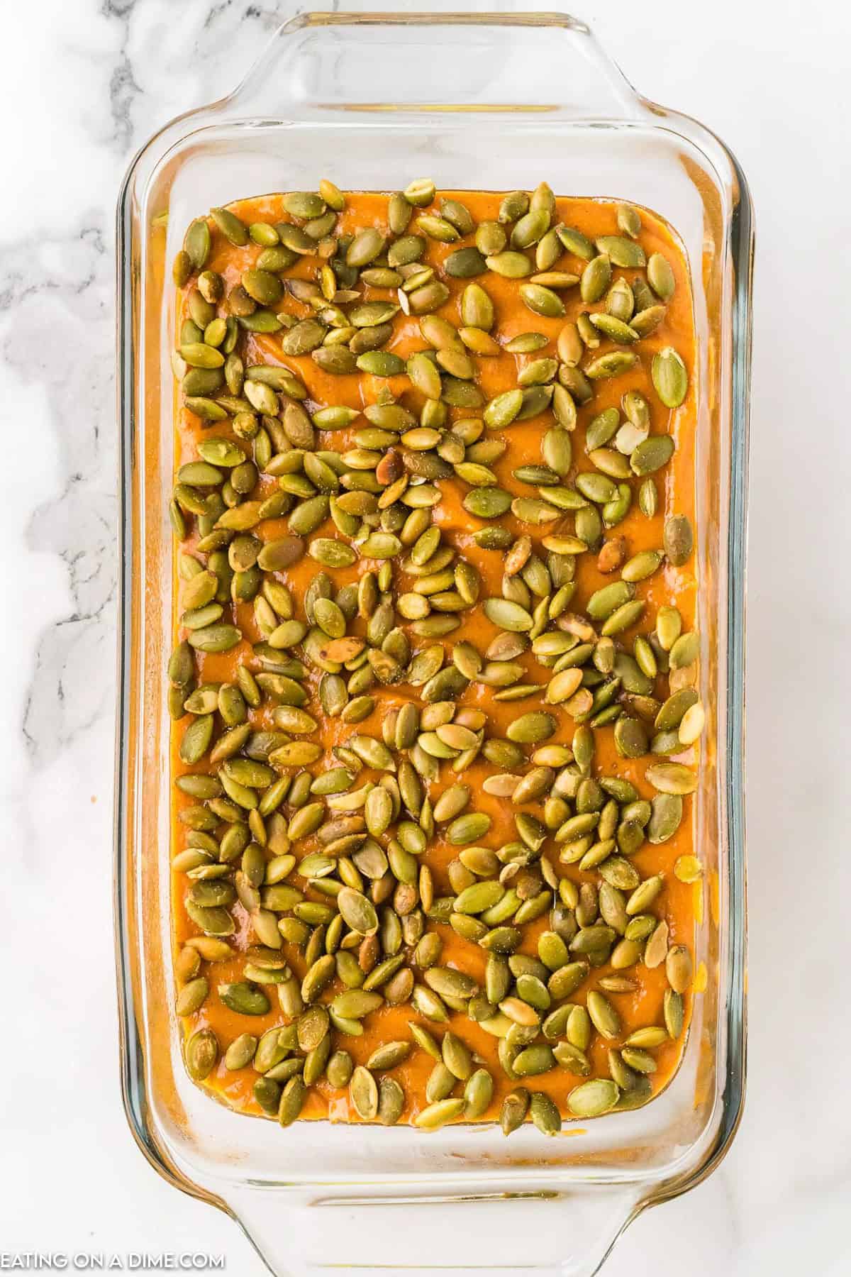 A glass baking dish filled with pumpkin bars reminiscent of a Starbucks Pumpkin Bread Recipe is topped with green pepitas. The vibrant orange of the bars contrasts beautifully with the seeds as the dish rests elegantly on a white marble surface.