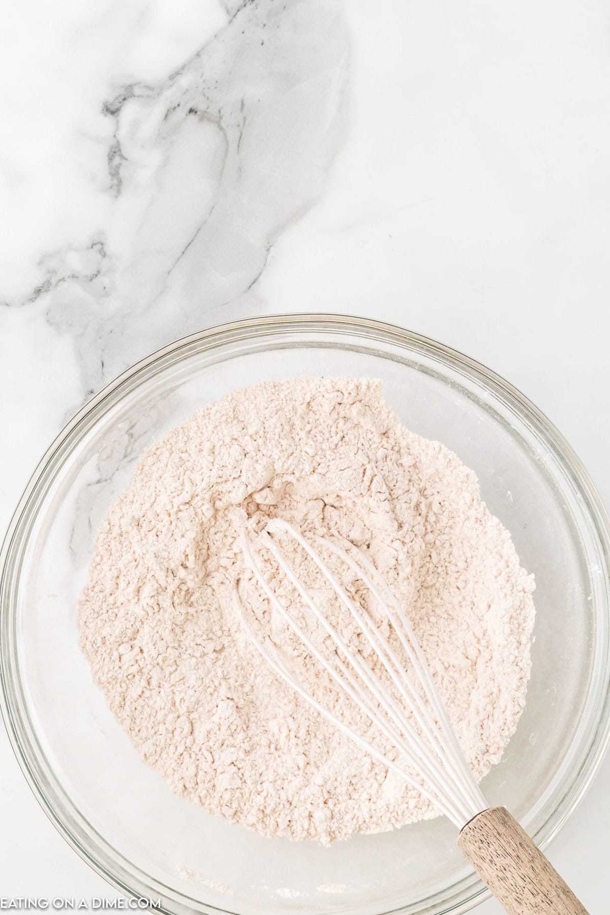 A glass bowl filled with a light brown flour mixture, reminiscent of a Starbucks Pumpkin Bread recipe, sits on a marble countertop. A whisk with a wooden handle rests inside the bowl, partially buried in the fragrant blend.