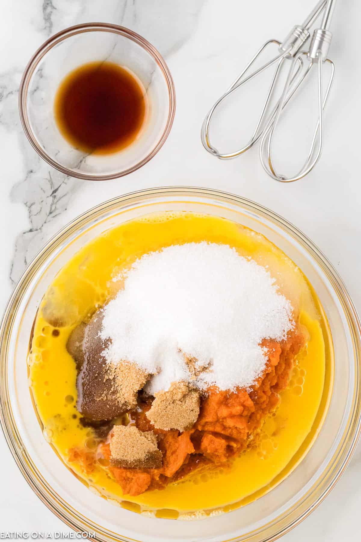 A glass bowl filled with pumpkin purée, sugar, brown sugar, and eggs evokes the beloved Starbucks Pumpkin Bread recipe. A small bowl of vanilla extract and metal beaters rest nearby on a marble surface, ready to bring the flavors together.