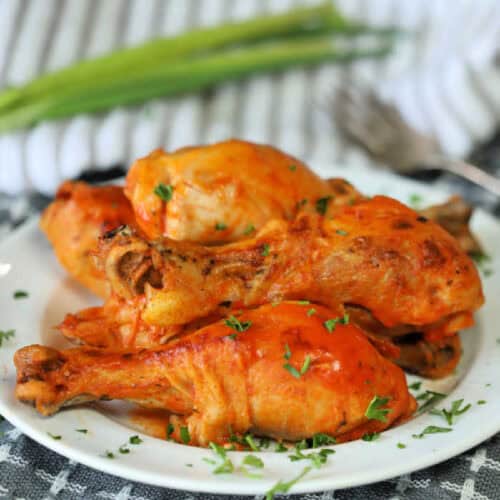 A white plate showcases Crock Pot Buffalo Chicken Drumsticks coated in a vibrant orange sauce, garnished with chopped parsley. A fork and a green onion rest nearby on a patterned cloth, hinting at the delicious flavors of this simple recipe.