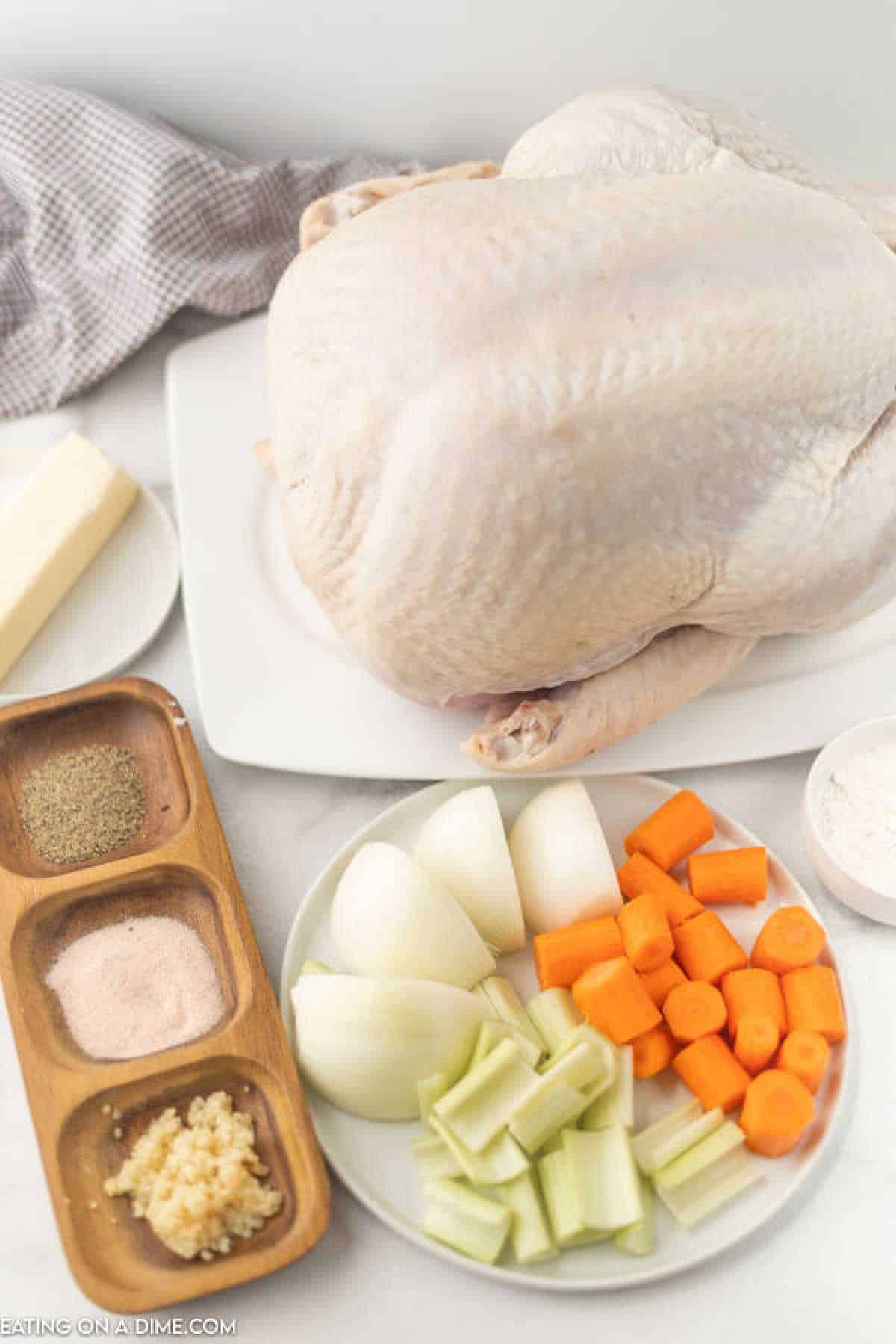 A raw turkey on a white plate is surrounded by ingredients like butter, garlic, chopped carrots, celery, onions, salt, pepper, and flour—perfect for those learning how to cook a turkey in a bag. A checkered cloth is partially visible in the background.