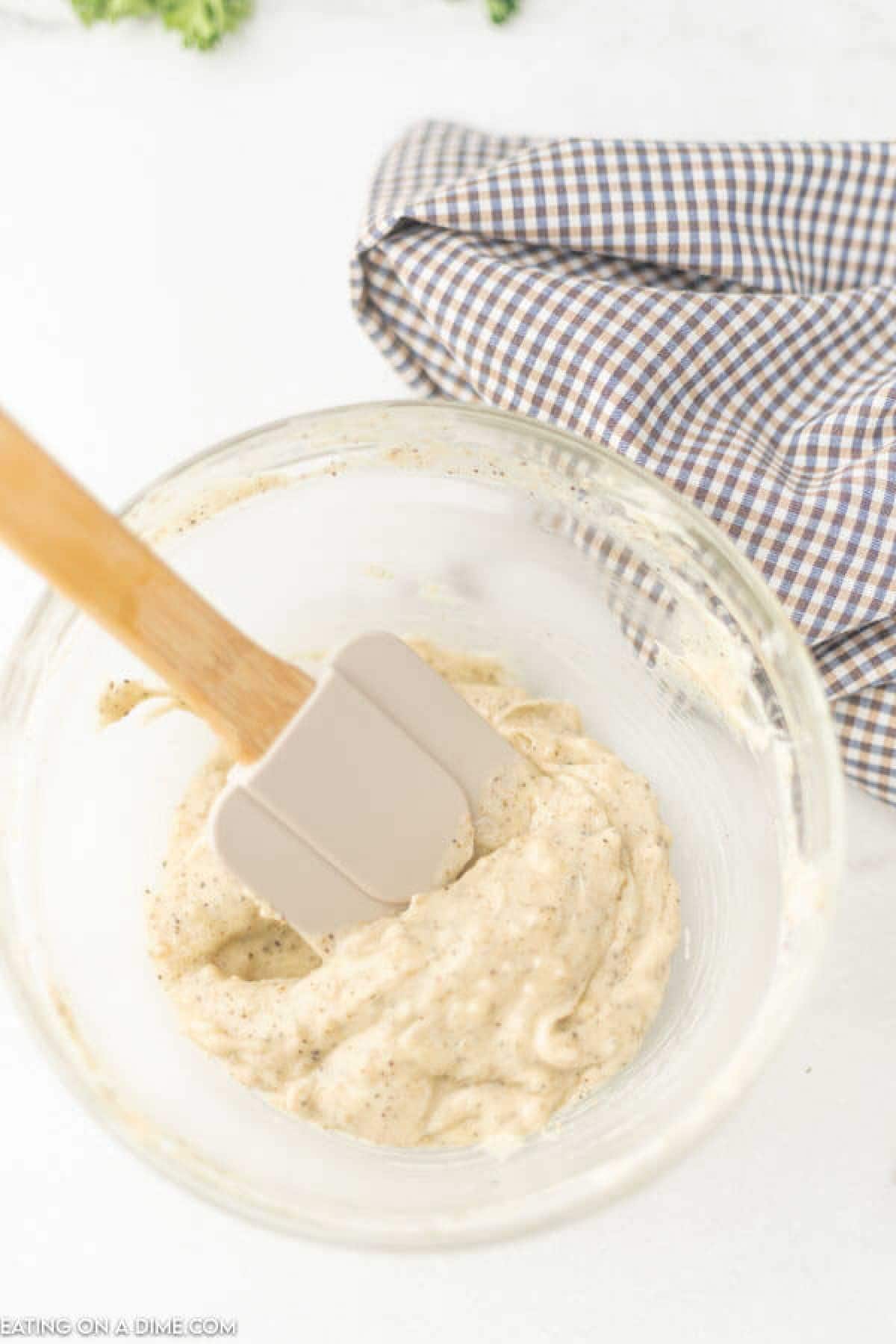 A glass bowl of creamy sauce, peppered to perfection, is being mixed by a grey spatula beside a checkered cloth on a light surface—an essential accompaniment when discovering how to cook a turkey in a bag.