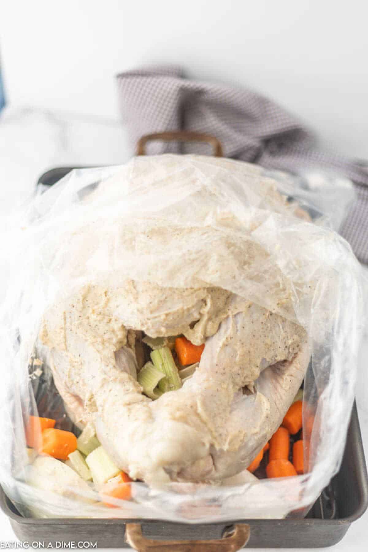 A raw turkey seasoned with a creamy blend is expertly placed in a roasting bag, nestled among chopped carrots and celery in a metal roasting pan. If you're wondering how to cook a turkey in a bag, this method ensures juicy perfection every time, with a checkered cloth providing quaint background charm.