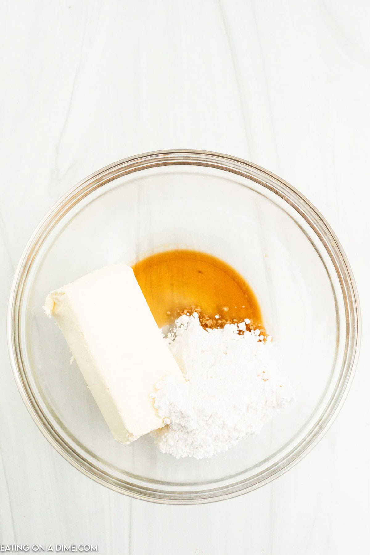 A glass bowl containing a stick of cream cheese, powdered sugar, and a small pool of vanilla extract, ready to be mixed for creating delightful Strawberry Santas.