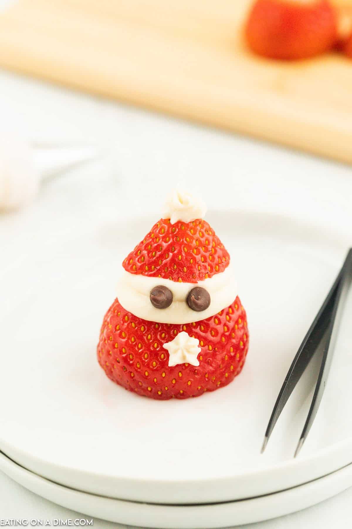 A delightful Strawberry Santa dessert sits on a white plate, with a dollop of cream as the hat and beard, and two small chocolate chips for eyes. A pair of tweezers is placed beside this charming creation.