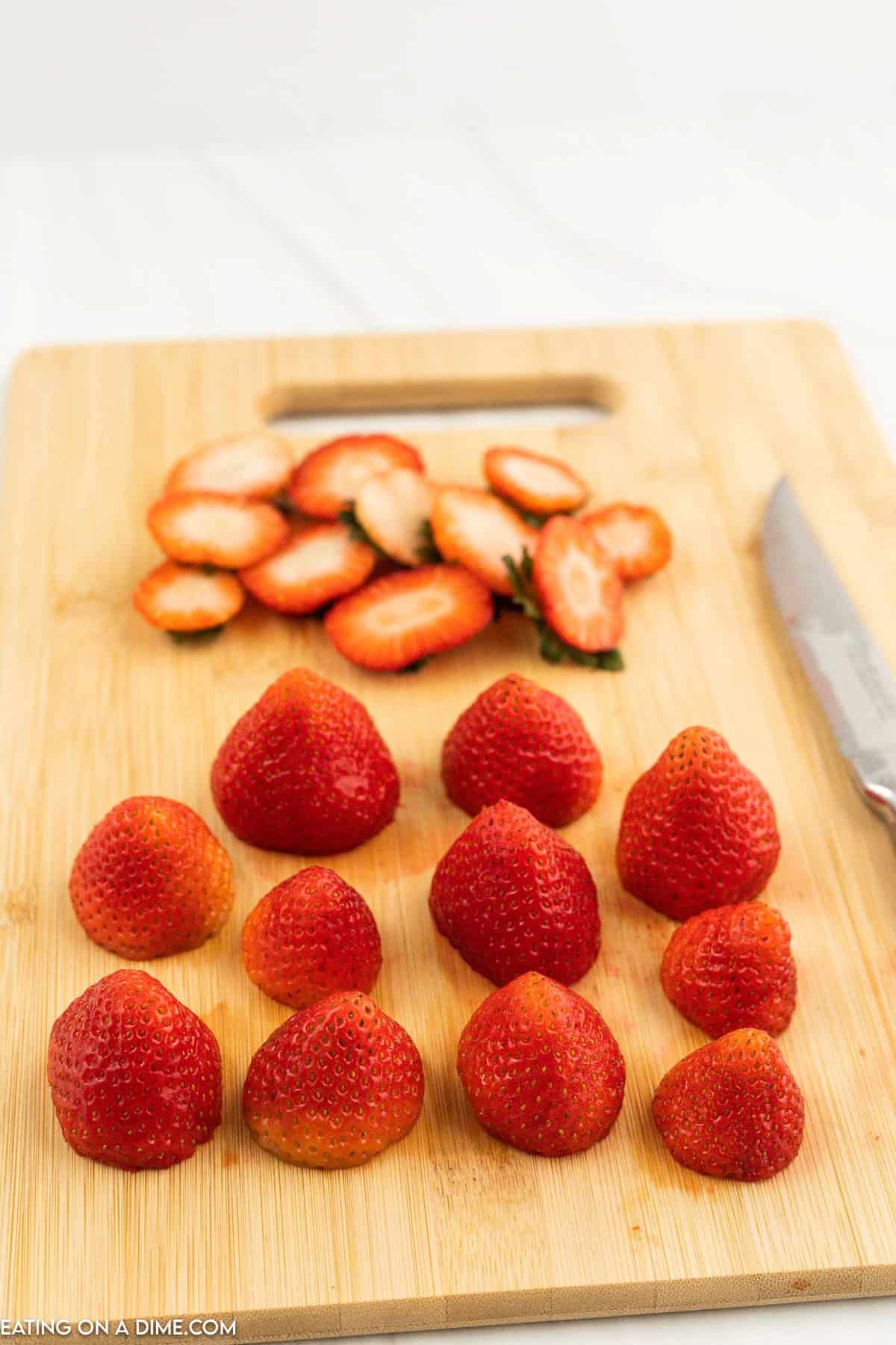 A wooden cutting board with a knife and several whole strawberries, some sliced with tops removed, resembling Strawberry Santas. The strawberries are evenly spaced, showing a preparation process for cooking or garnishing.