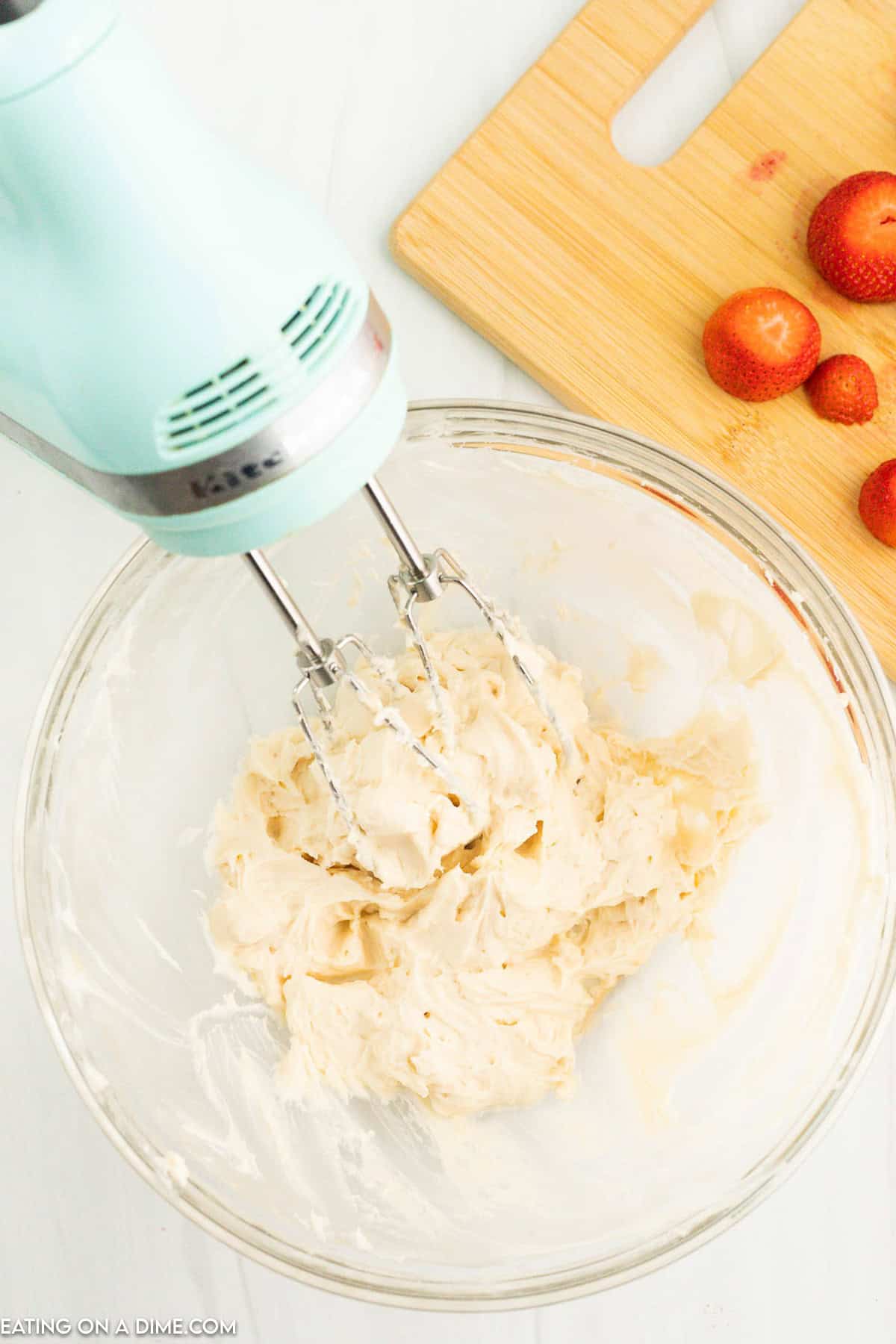 A turquoise electric mixer is blending a creamy mixture in a glass bowl. Beside the bowl, on a wooden cutting board, are some strawberries, some whole and some with tops removed. The scene suggests preparation of Strawberry Santas for a delightful holiday dessert.