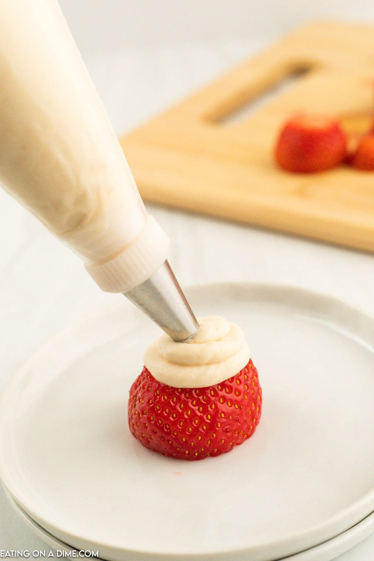 A piping bag with a stainless steel tip is used to pipe fluffy white cream onto a halved strawberry that sits on a white plate, creating adorable Strawberry Santas. The blurred background shows other halved strawberries on a wooden cutting board.