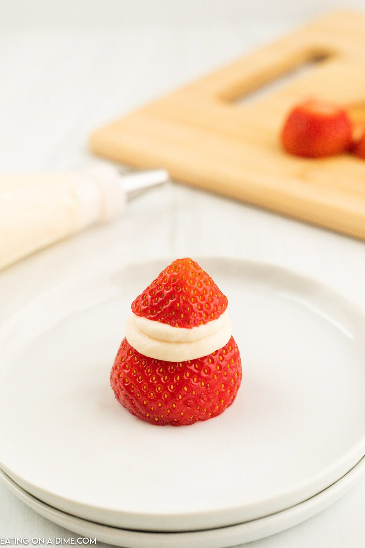 A strawberry on a white plate is sliced in the middle and filled with cream, resembling a mini Santa hat. In the background, another sliced strawberry and a piping bag with cream are on a wooden cutting board, creating an adorable scene of Strawberry Santas.