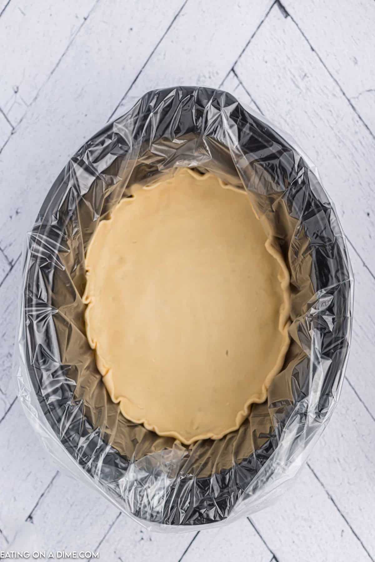 A slow cooker lined with plastic, filled with creamy brown batter destined for a Crock Pot Pecan Pie, sits on a white wooden herringbone floor. The image shows a top-down view of the prepared mixture, ready for cooking.