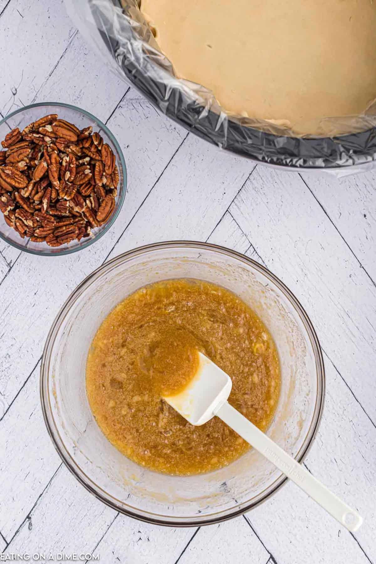 A bowl of mixed ingredients and a white spatula sits on a light wooden surface, ready to create the perfect Crock Pot Pecan Pie. Nearby, a small bowl of pecans waits beside a baking dish with dough covered in plastic wrap, promising homemade goodness.