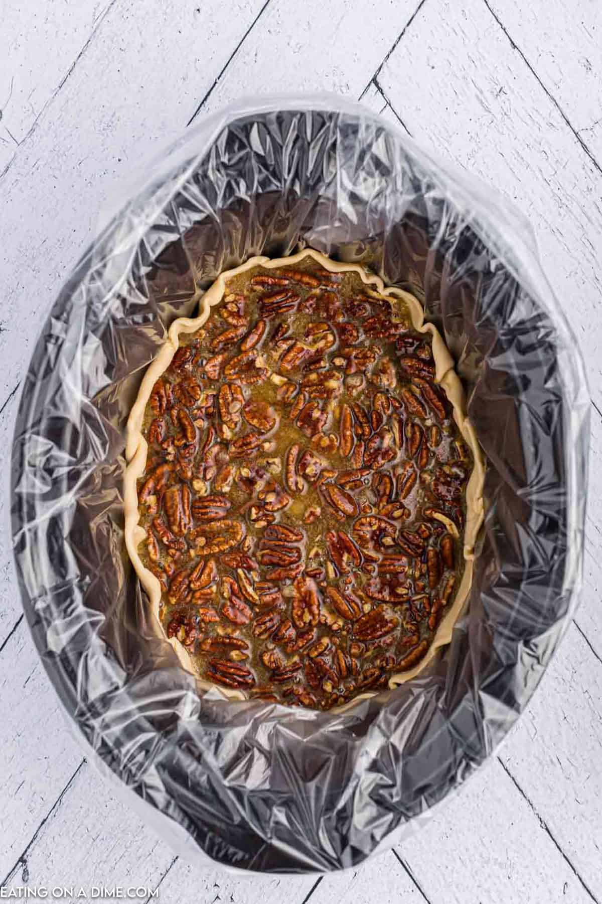 A Crock Pot Pecan Pie is baking inside a slow cooker lined with a transparent plastic bag. The pie features a golden crust, topped with glazed pecans, as it simmers to perfection on a white wooden surface.