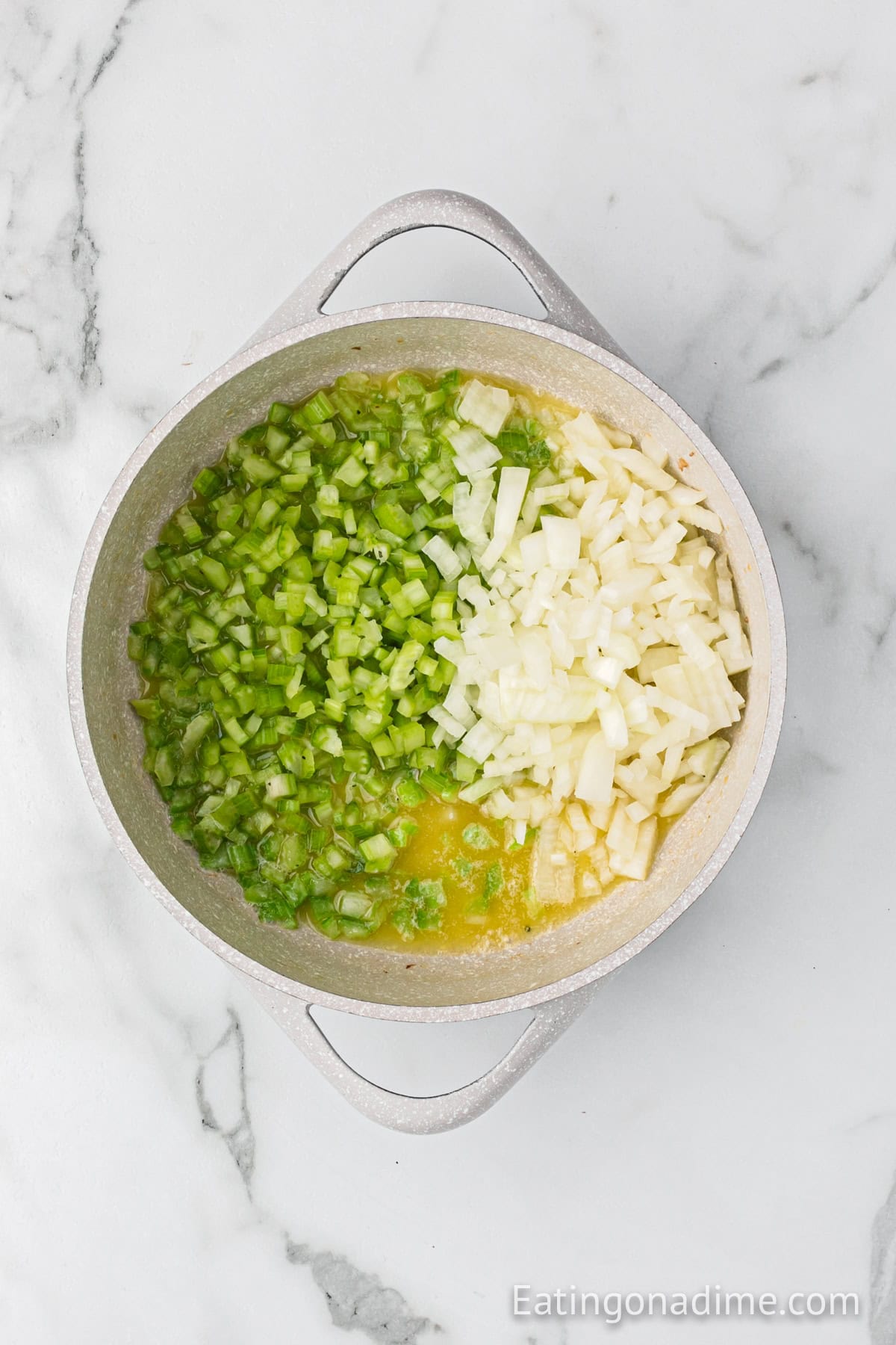 Overhead view of a pot containing diced green peppers and chopped onions sautéing in melted butter, a perfect start for cornbread sausage stuffing. The pot is placed on a white marble surface.