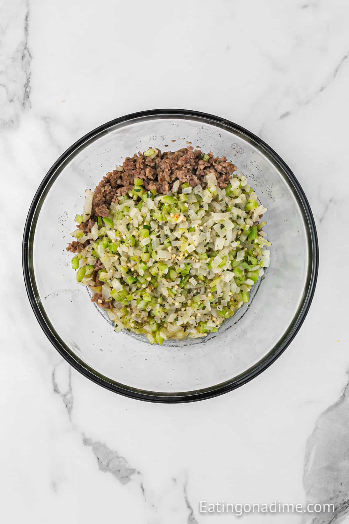 A glass bowl containing a mixture of cooked minced beef, chopped onions, celery, and spices sits on a white marble surface. Reminiscent of a hearty cornbread sausage stuffing recipe, these ingredients are perfectly blended to create a savory delight.