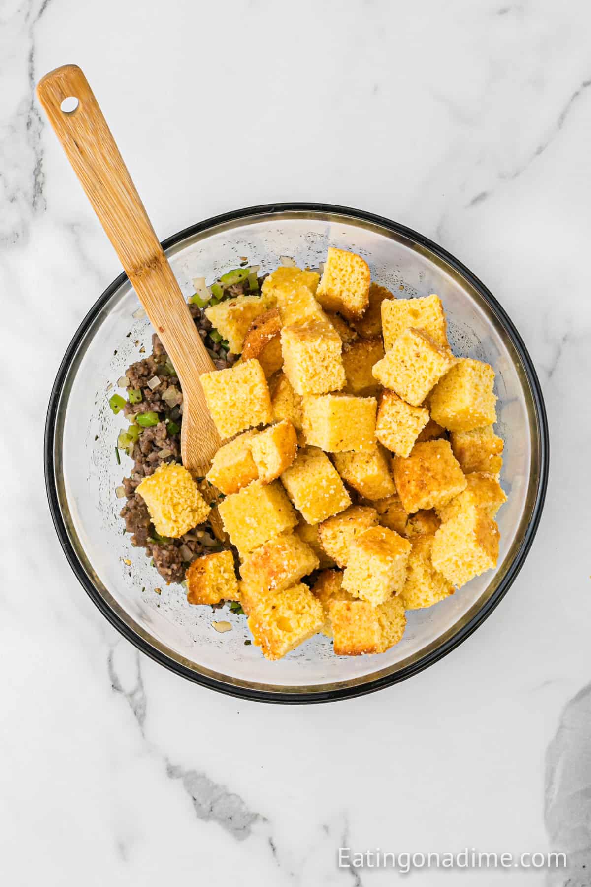 Sausage mixture in a bowl topped with cubed cornbread with a wooden spoon